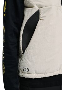 Beige jas met zwarte, met een rits afsluitbare mesh-zijzak en een zwarte "223"-print vlak bij de zoom, gedragen over een zwart shirt met lange mouwen met gele tekst.