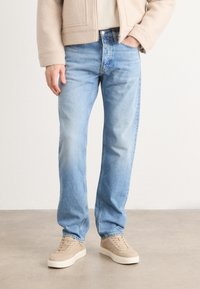Jeans de denim azul claro con corte recto, que presentan desvanecimiento y una sutil textura. Combinados con zapatillas beige y una blusa crema.