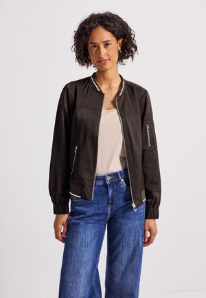 Woman with short curly hair wearing black bomber jacket, beige top, and blue jeans standing against plain background.