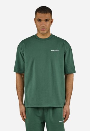 Mann iført mørkegrønn Pegador T-skjorte og matchende bukser, vendt fremover med nøytral uttrykk mot ensfarget lys bakgrunn.