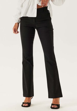 Kvinde iført sorte højtaljede bukser og sorte åbne hæle, kombineret med en hvid langærmet cropped top, mod en ensfarvet baggrund.