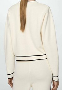 Persona con lunghi capelli castani che indossa un completo di maglione a maglia color crema e pantaloni, con bordi neri su polsini, orlo e vita, mostrata di spalle.