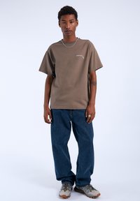 Camiseta de algodón marrón de manga corta con un pequeño gráfico, combinada con jeans azules y zapatillas grises con acentos verdes y detalles texturizados.