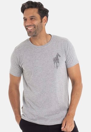 Homme souriant avec des cheveux foncés portant un T-shirt gris clair avec un design géométrique de cheval sur la poitrine gauche, se tenant avec une main dans la poche.