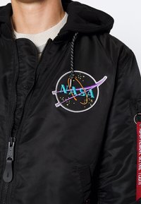 Czarna kurtka typu bomber z kapturem, z wyszywanym logo NASA w kolorowych nićmi. Posiada zamek błyskawiczny oraz czerwoną metkę z tkaniny na rękawie.