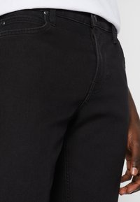 Black denim jeans feature a fitted design, front pockets with stitching details, and a smooth texture, shown at the hip and thigh area.