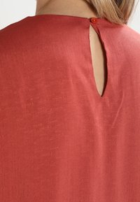 Červený texturovaný top s klíčovým otvorem na zádech, který je zajištěn kontrastním knoflíkem. Tkanina má jemný lesk.