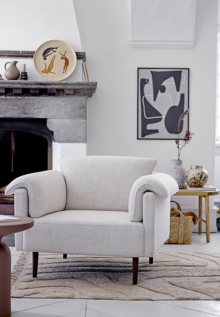 Fauteuil rembourré gris clair avec accoudoirs arrondis et pieds en bois, posé sur un tapis à motifs, entouré d'objets décoratifs.