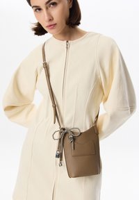 Bolso cruzado de cuero marrón con superficie texturizada, correa ajustable y detalle de cadena decorativa. Colocado sobre un vestido color crema.