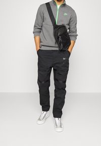 Sudadera de forro polar gris con detalles verdes, combinada con pantalones cargo negros que tienen múltiples bolsillos y zapatillas negras con suelas blancas.