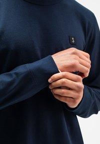 Dunkelblauses Langarmhemd aus glattem Baumwollstoff mit Rundhalsausschnitt und einem kleinen schwarzen Etikett mit einem weißen Logo auf der Brust.