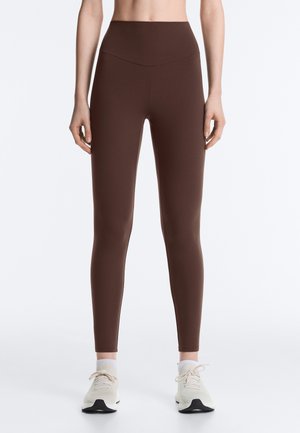 Femme portant des leggings bruns taille haute et des baskets blanches, debout devant un fond clair uni.