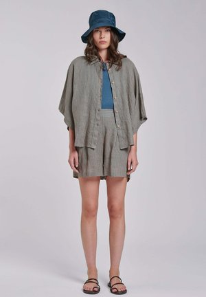 Frau mit blauem Fischerhut, blauem Top, lockerem kurzärmligem gestreiften Hemd zum Knöpfen, passenden Shorts und schwarzen Sandalen, stehend vor schlichtem Hintergrund.