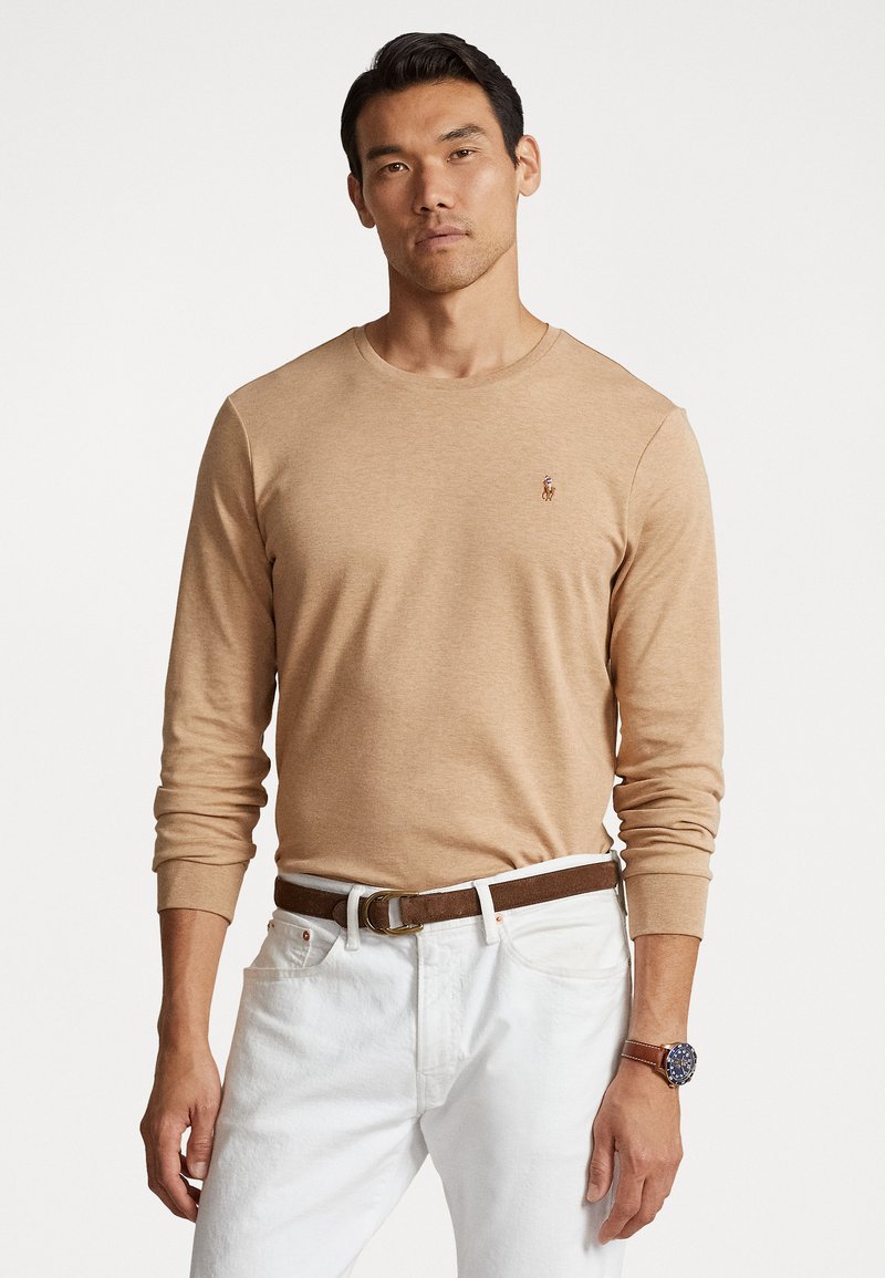 Lichtbruin shirt met lange mouwen, gemaakt van een zachte stof, met een ronde halslijn en een klein logo op de borst, gecombineerd met witte jeans en een brown riem.