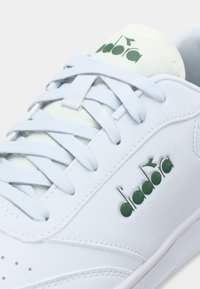 Άσπρο δερμάτινο αθλητικό παπούτσι με ανοιχτό γαλάζια κορδόνια, πράσινες λογότυπες λεπτομέρειες και υφασμένο επάνω μέρος. Διαθέτει οπές αερισμού και λεία λαστιχένια σόλα.