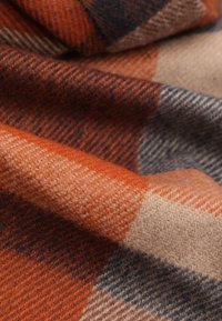 Wełniany koc w kratę w kolorach pomarańczowym, beżowym i ciemnoniebieskim. Tekstura wygląda na miękką i ciepłą, z tkanym wykończeniem.