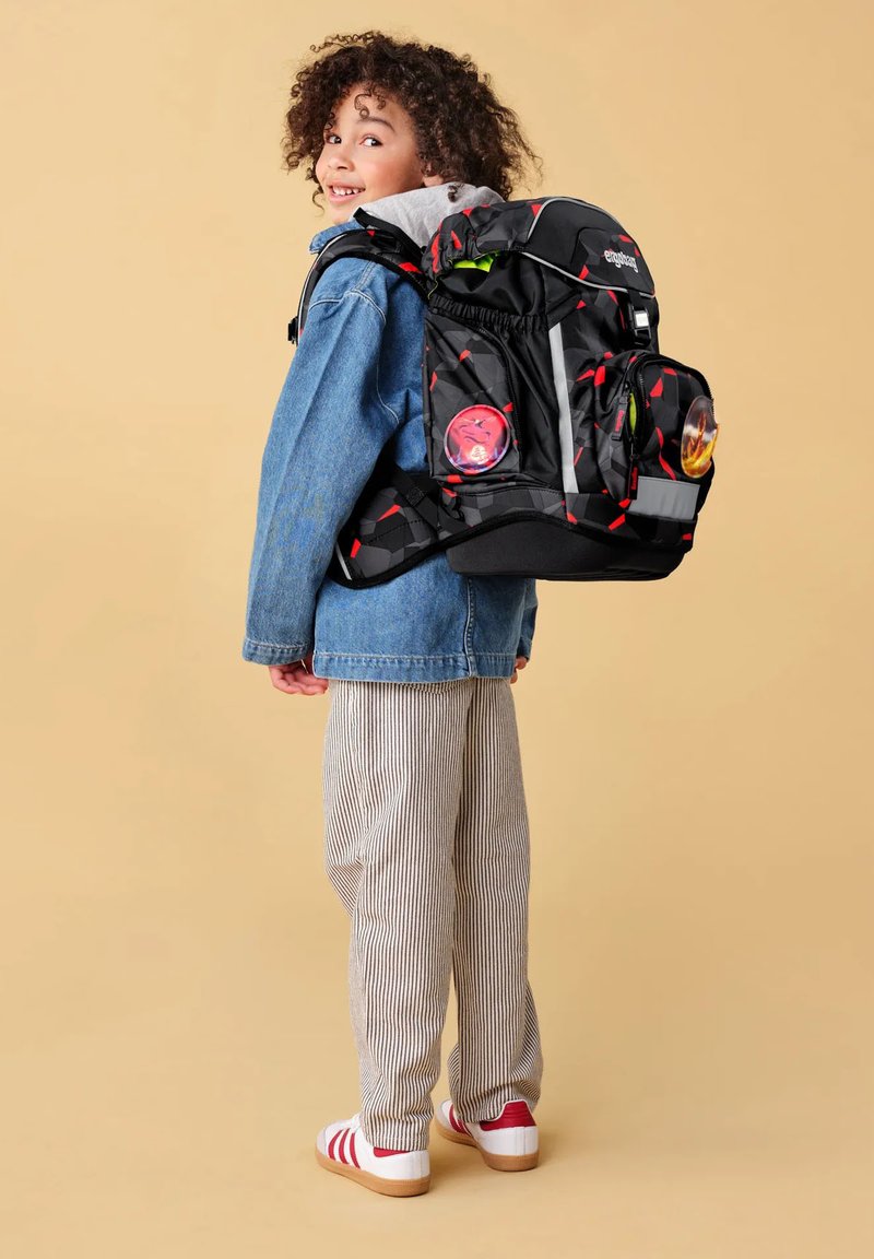 Kind mit lockigem Haar, das eine Jeansjacke und gestreifte Hosen trägt, lächelt mit großem schwarz-rotem Rucksack vor beigem Hintergrund.