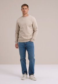 Bežs kreklā ar apkakli, izgatavots no mīksta auduma, apvienots ar gaišiem ziliem džinsiem un baltām sporta kurpēm. Modelis stāv uz neitrāla fona.