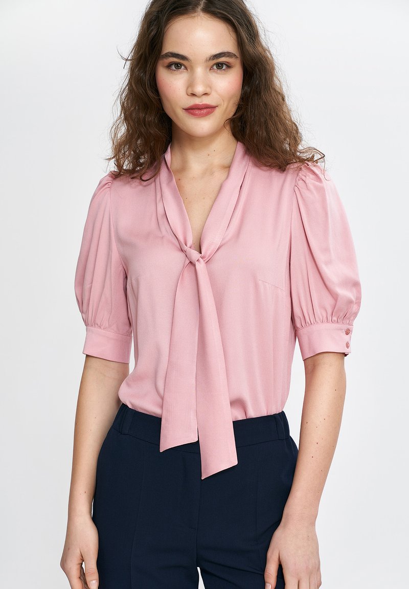 Jasnoróżowa bluzka z krótkim rękawem, z wiązanym dekoltem, falbankowymi rękawami i guzikami przy mankietach; gładka faktura materiału.