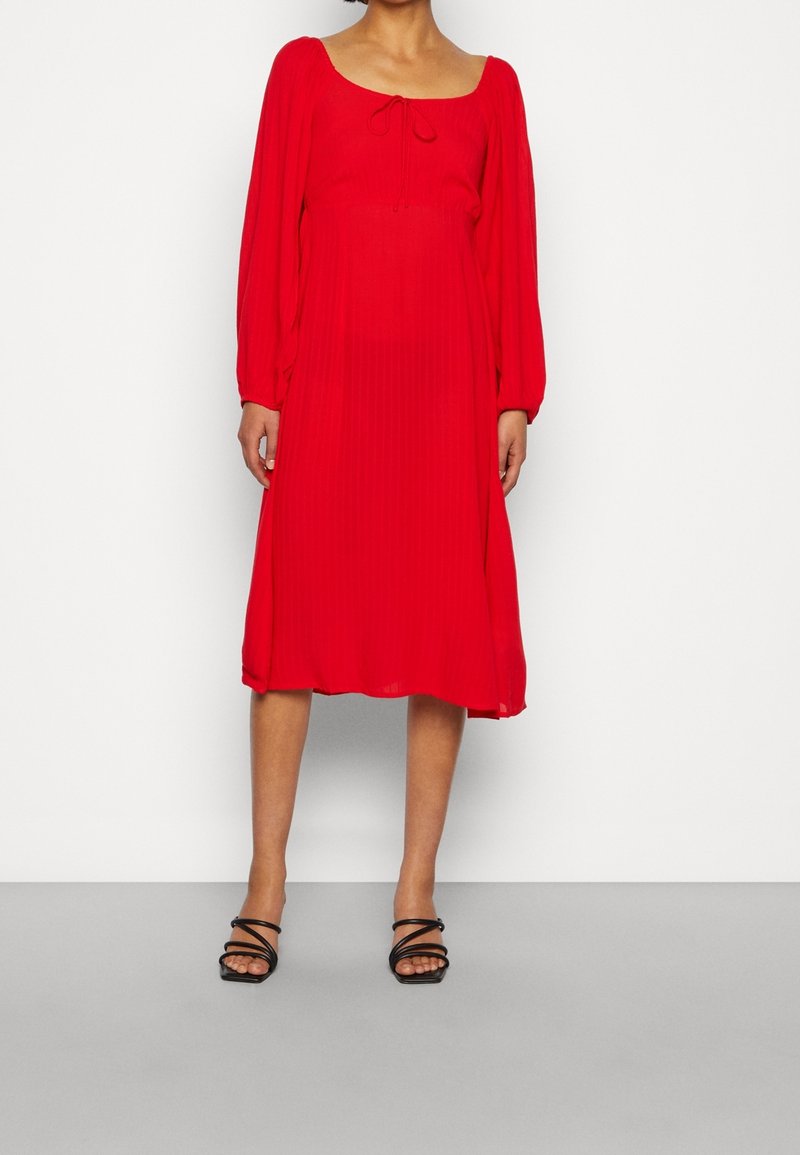 Červené plisované midi šaty se čtvercovým výstřihem, dlouhými rukávy a vázáním vpředu. Vpředu doplněné tenkými černými sandály na špagetových řemíncích.