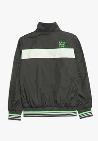 Chaqueta ligera negra con una franja horizontal blanca en la parte trasera, detalles verdes y el texto "CARS JEANS 1982" en la parte superior izquierda.
