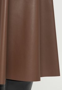 Brown faux leather skirt with a smooth texture, flaring from the waist. The hem is straight and sits just above black leggings with a shiny finish.