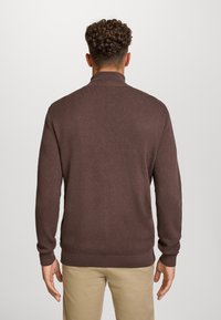 Brun strikkegenser med høy krage og ribbestrikkede ermer. Har et strukturert mønster, tettsittende silhuett, kombinert med lyse beige bukser.