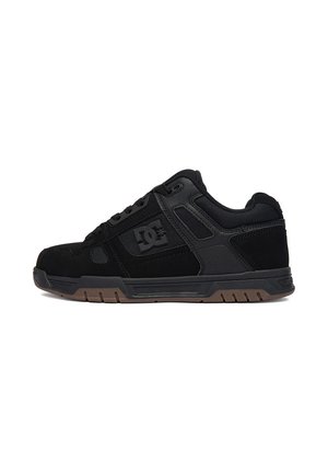 Zapatilla negra de corte bajo con diseño de cordones, cuello acolchado y suela gruesa con el logo de DC en el lateral y suela exterior marrón texturizada.