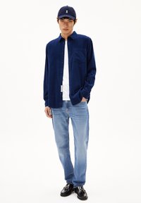 Marineblauwe overhem shirt met een zak, witte t-shirt eronder, lichtblauwe rechte spijkerbroek en zwarte schoenen. Donkerblauw pet met logo.