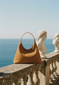 Καφέ δερμάτινη τσάντα που ξεκουράζεται πάνω σε μια πέτρινη μπαλustrάδα με κλασικά αγάλματα προτομών και θέα στη θάλασσα στο φόντο.