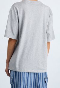 Persona che indossa una maglietta grigia chiara e larga e pantaloni blu a righe con tasche laterali, mostrata di spalle su uno sfondo chiaro e semplice.