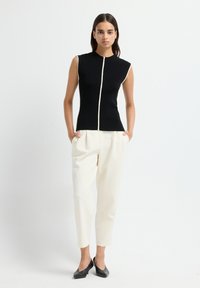 Donna che indossa un top senza maniche nero con rifiniture bianche, pantaloni bianchi a vita alta e gamba affusolata, e scarpe nere con tacco basso, in piedi con le mani nelle tasche.