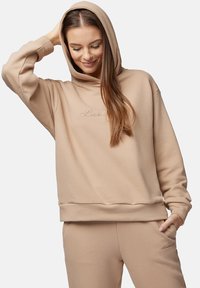 Beżowa bluza z kapturem, wykonana z miękkiego materiału, o luźnym kroju, z wiązanym kapturem i haftowanym logotypem z przodu. Połączona z pasującymi spodniami.