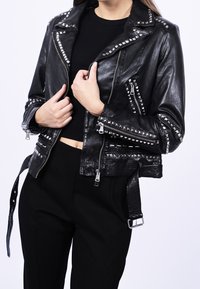 Veste de motard en cuir noir avec des clous argentés, des poches à fermeture éclair et une taille ceinturée. Caractérisée par un style court et une finition texturée.