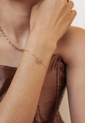 Feines Armband aus Roségoldkette mit einem Lebensbaum-Anhänger, getragen am erhobenen Handgelenk, kombiniert mit einer passenden Halskette und einem braunen Satintop.