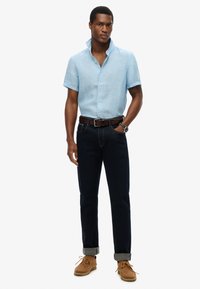 Maglietta azzurra chiara a maniche corte con bottoni, jeans in denim scuro con risvolti, cintura in pelle marrone e scarpe in pelle beige con lacci.