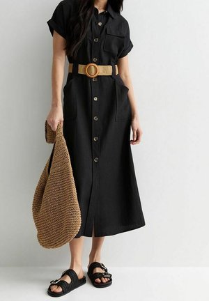 Žena v černých midi šatech s knoflíky a založenými rukávy, s opáleným pásem, drží velkou opálenou pletenou tašku, má na nohou černé sandály s přezkami.