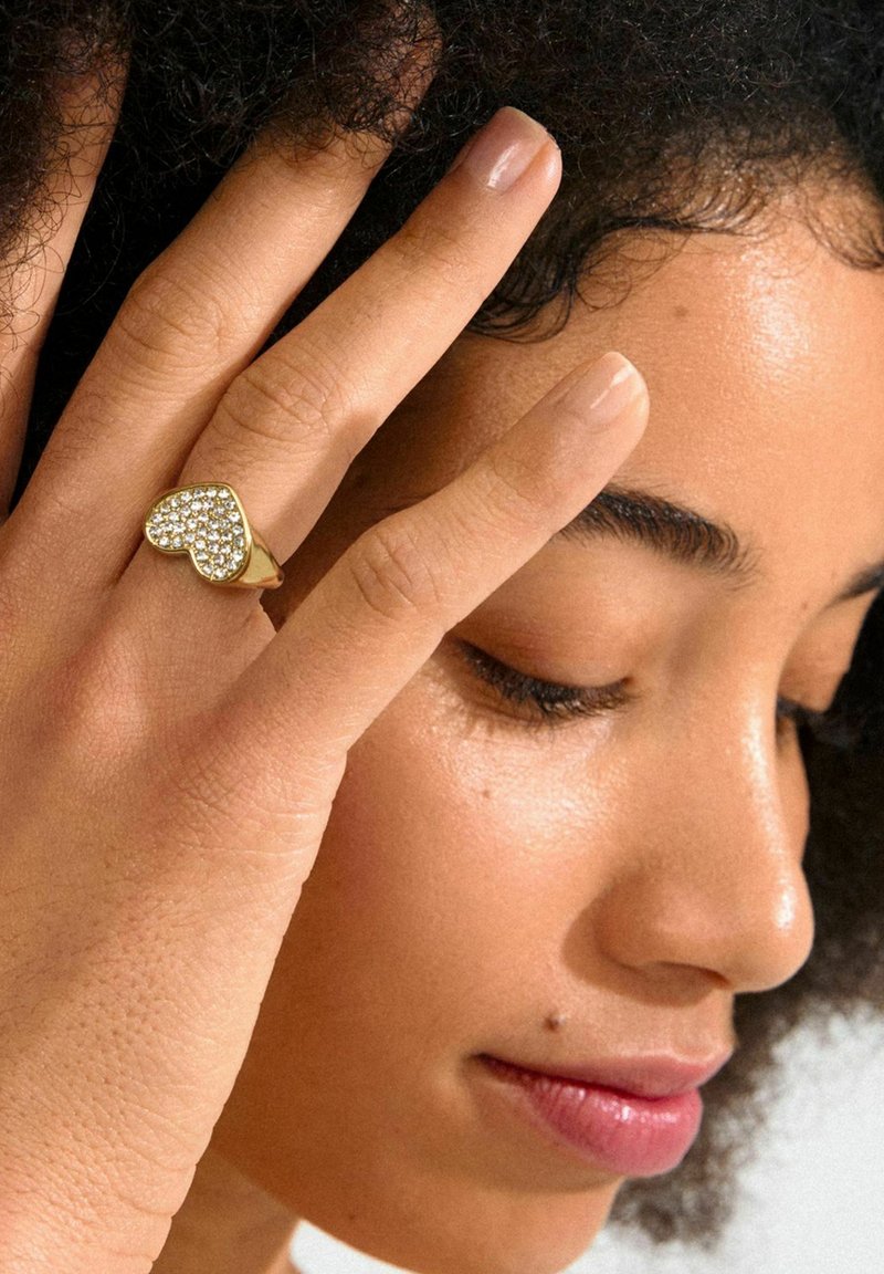 Anillo dorado en forma de corazón con acentos de cristal, con un acabado pulido. Los dedos muestran uñas naturales contra una piel texturizada.