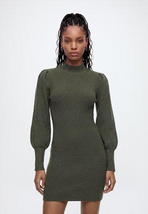 Femme aux longs cheveux tressés portant des boucles d’oreilles créoles dorées et une robe en maille côtelée vert olive avec des manches longues bouffantes.
