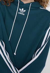 Teal huvtröja med vita ränder längs ärmarna och vita dragsnören; har Adidas-logotyp på bröstet i vit brodering.