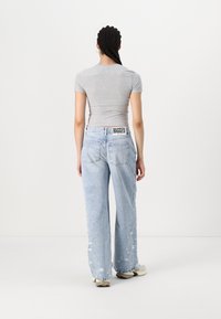 Jeans de tiro alto en azul claro de pierna ancha con detalles desgastados y una etiqueta negra "RAGGED" en la cintura, combinados con una camiseta ceñida gris.
