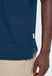 Nærbilde av en person som har på seg en marineblå skjorte med en liten blandingsetikett og beige bukser, hånden avslappet ved siden av.