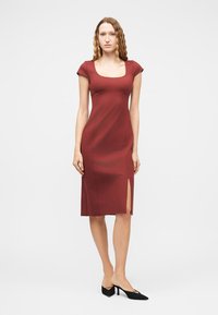 Γυναίκα με κυματιστά μαλλιά που φοράει εφαρμοστό μπορντό φόρεμα με κοντά μανίκια και σκίσιμο στο πλάι, συνδυασμένο με μαύρες μυτερές γόβες.