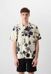 Camicia a maniche corte con bottoni in tessuto giallo chiaro con stampa floreale nera. Presenta un colletto e una tasca sul petto. Combinata con pantaloni corti neri.