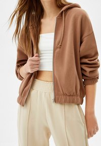 Kvinna som bär en brun zip-up hoodie över en vit crop top och krämfärgade högmidjade byxor med elastisk midja.