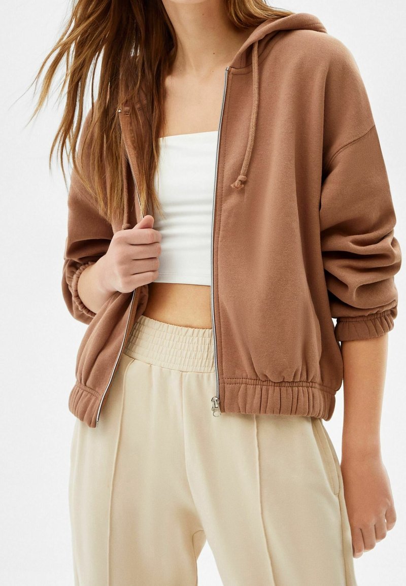 Žena v hnědé mikině na zip přes bílý crop top a krémových kalhotách s vysokým pasem a elastickým pasem.