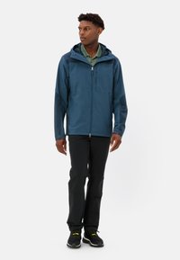 Giacca impermeabile blu con cappuccio, zip frontale e tasche laterali, abbinata a pantaloni neri e scarpe sportive. Texture liscia e vestibilità moderna.