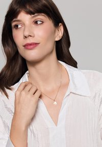 Złoty naszyjnik z wisiorkiem na delikatnym łańcuszku, z okrągłym wisiorkiem o kwiatowym wzorze, noszony na białej, teksturowanej bluzce.