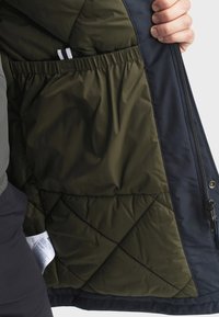 Unutrašnjost paketne jakne u maslinastoj zelenoj boji s dijamantnim uzorkom. Sadrži podstavu u tamnoplavoj boji, patentni zatvarač i elastične otvore za džepove.