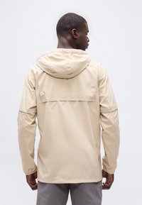 Άνδρας που φοράει μπεζ μπουφάν με κουκούλα και γκρι παντελόνι, στέκεται με την πλάτη γυρισμένη σε απλό λευκό φόντο.